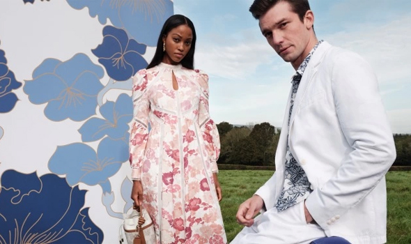 a man modelling a white suit and a woman modelling a white dress with pink flowers in a field